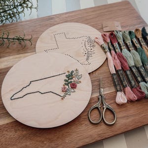May include: Wooden embroidery hoops with state outlines, one featuring floral embroidery. Includes embroidery floss in various colors and small scissors. The items are arranged on a wooden cutting board.