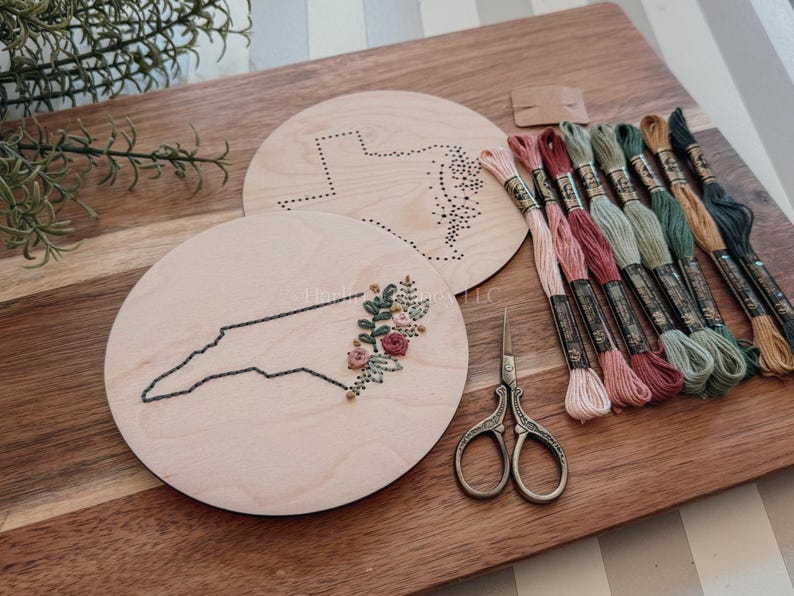 May include: Wooden embroidery hoops with state outlines, one featuring floral embroidery. Includes embroidery floss in various colors and small scissors. The items are arranged on a wooden cutting board.