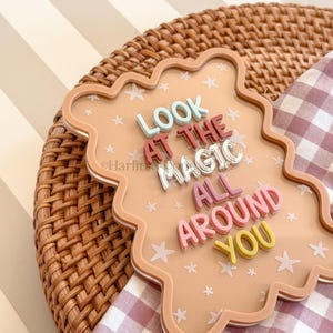 May include: A decorative, light brown, cloud-shaped sign with the words "LOOK AT THE MAGIC ALL AROUND YOU" in colorful, raised lettering. The sign is decorated with white star shapes and rests on a woven placemat and a checkered cloth.