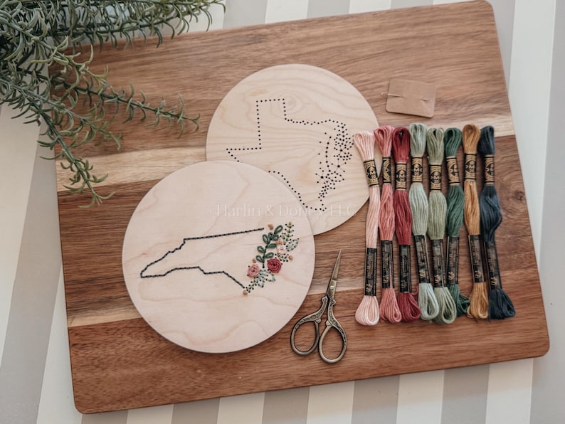 May include: Wooden embroidery hoops featuring state outlines, one with a floral design. A collection of colorful embroidery floss, small scissors, and a blank tag are arranged on a wooden cutting board. The hoops are approximately 6 inches in diameter.