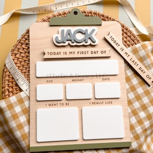 May include: A wooden clipboard sign with the name JACK in raised letters. It features text reading TODAY IS MY FIRST DAY OF and TODAY IS MY LAST DAY OF, with blank white rectangular spaces for age, height, date, goals, and interests.