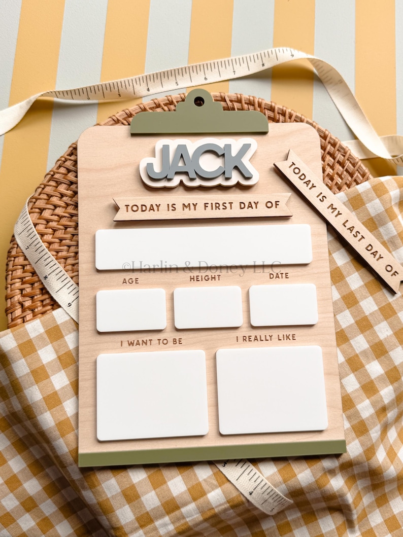 May include: A wooden clipboard sign with the name JACK in raised letters. It features text reading TODAY IS MY FIRST DAY OF and TODAY IS MY LAST DAY OF, with blank white rectangular spaces for age, height, date, goals, and interests.