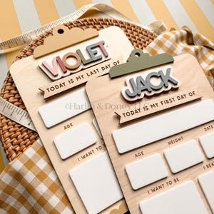 May include: Two wooden clipboard-style signs with white dry-erase sections for recording milestones. One sign reads VIOLET and TODAY IS MY LAST DAY OF, while the other reads JACK and TODAY IS MY FIRST DAY OF. Both feature blank fields for age, height, date, and goals.