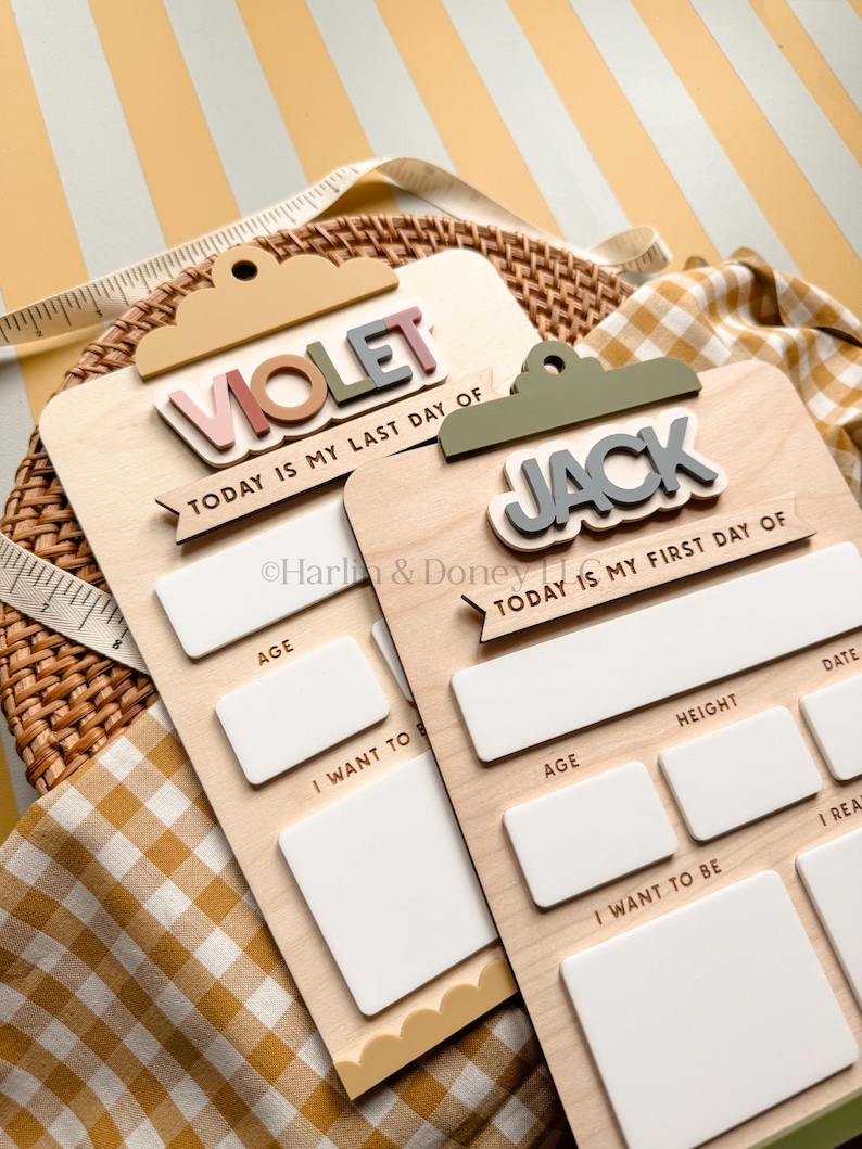 May include: Two wooden clipboard-style signs with white dry-erase sections for recording milestones. One sign reads VIOLET and TODAY IS MY LAST DAY OF, while the other reads JACK and TODAY IS MY FIRST DAY OF. Both feature blank fields for age, height, date, and goals.