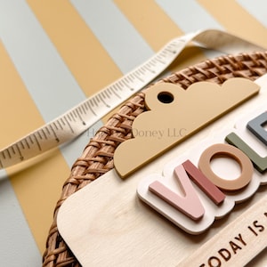 May include: A circular woven rattan wall plaque featuring a light wood base with a cloud-shaped topper. The plaque displays the name VIOLET in raised, multi-colored letters and the text TODAY IS. A white measuring tape shows a width of 3 inches.