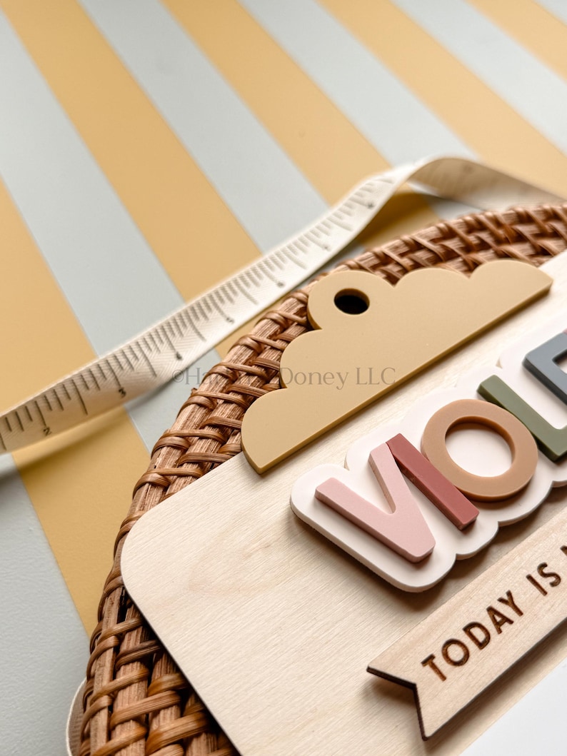 May include: A circular woven rattan wall plaque featuring a light wood base with a cloud-shaped topper. The plaque displays the name VIOLET in raised, multi-colored letters and the text TODAY IS. A white measuring tape shows a width of 3 inches.