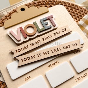 May include: A wooden milestone board featuring the name VIOLET in multi-colored 3D letters at the top. It includes two wooden banners reading TODAY IS MY FIRST DAY OF and TODAY IS MY LAST DAY OF, with white rectangular spaces for age, height, and goals.
