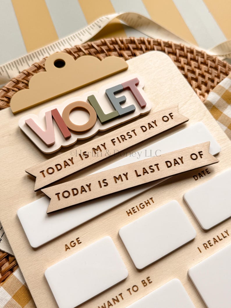 May include: A wooden milestone board featuring the name VIOLET in multi-colored 3D letters at the top. It includes two wooden banners reading TODAY IS MY FIRST DAY OF and TODAY IS MY LAST DAY OF, with white rectangular spaces for age, height, and goals.