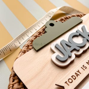 May include: A wooden plaque featuring a green clip top, the name JACK in raised gray letters on a white background, and the text TODAY IS M. A white measuring tape shows a length of approximately 6 inches along the top edge of the plaque.