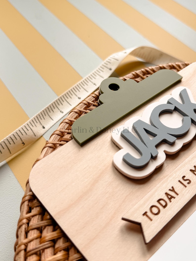 May include: A wooden plaque featuring a green clip top, the name JACK in raised gray letters on a white background, and the text TODAY IS M. A white measuring tape shows a length of approximately 6 inches along the top edge of the plaque.