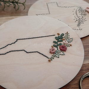 May include: Two round wooden embroidery hoops. One features a stitched outline of North Carolina with floral embroidery. The other has a dotted outline of a state. Embroidery floss and scissors are also visible.