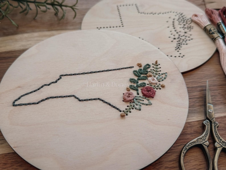 May include: Two round wooden embroidery hoops. One features a stitched outline of North Carolina with floral embroidery. The other has a dotted outline of a state. Embroidery floss and scissors are also visible.