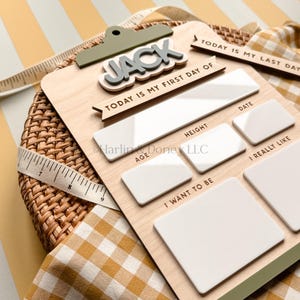 May include: A wooden milestone board featuring the name JACK in raised grey letters, with white acrylic sections for writing details. Text includes TODAY IS MY FIRST DAY OF, DATE, HEIGHT, AGE, I REALLY LIKE, and I WANT TO BE. A measuring tape is visible.