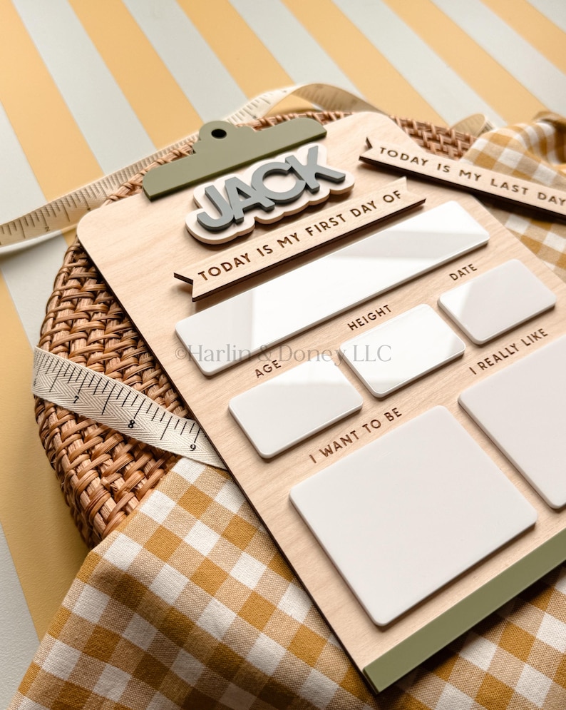May include: A wooden milestone board featuring the name JACK in raised grey letters, with white acrylic sections for writing details. Text includes TODAY IS MY FIRST DAY OF, DATE, HEIGHT, AGE, I REALLY LIKE, and I WANT TO BE. A measuring tape is visible.