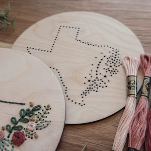 May include: Two unfinished wooden embroidery hoops with a Texas outline and floral design. Several skeins of embroidery floss in shades of pink, red, and green are arranged nearby, along with a needle. The hoops are on a wooden surface.