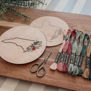 May include: Wooden embroidery hoops featuring state outlines, with floral embroidery on one. A collection of colorful embroidery floss, scissors, and a wooden cutting board complete the craft setup. The hoops are approximately 6 inches in diameter.