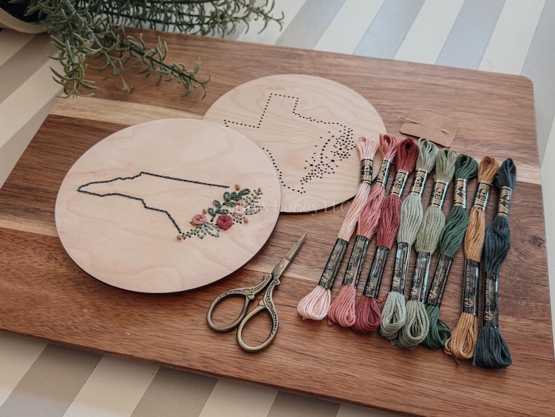 May include: Wooden embroidery hoops featuring state outlines, with floral embroidery on one. A collection of colorful embroidery floss, scissors, and a wooden cutting board complete the craft setup. The hoops are approximately 6 inches in diameter.