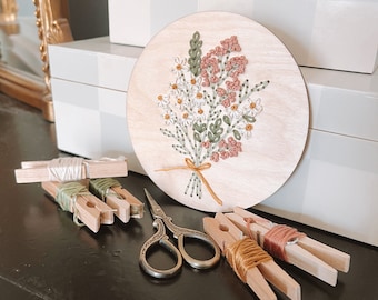 May include: A round wooden embroidery hoop featuring a floral design. The design includes white, pink, and green flowers. The hoop is surrounded by wooden pegs, scissors, and reels of thread.