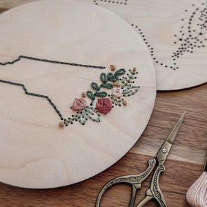 May include: Two wooden embroidery hoops, one featuring a state outline with floral embroidery. The other hoop has a dotted design. Pink and peach embroidery floss and small scissors are also visible.