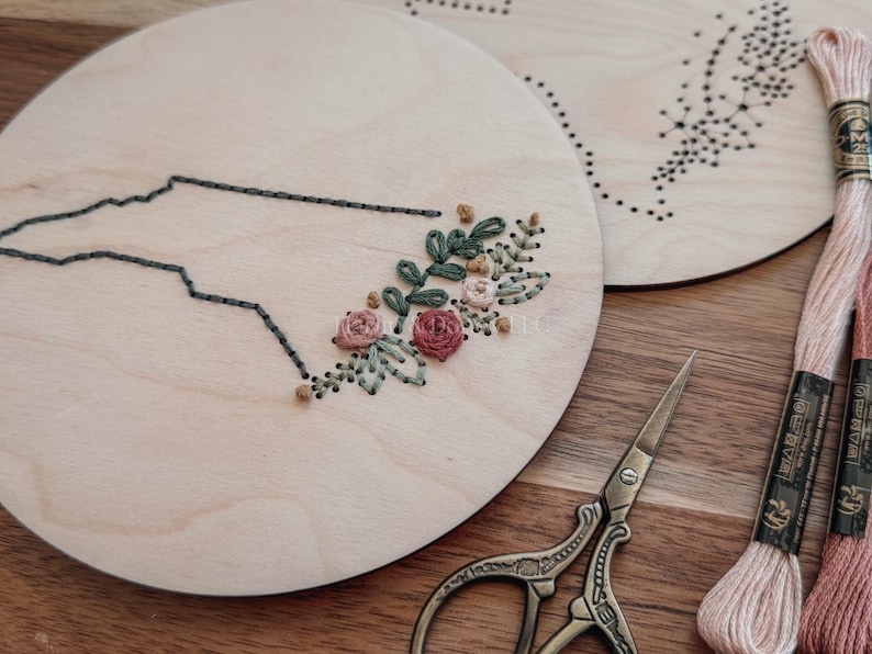 May include: Two wooden embroidery hoops, one featuring a state outline with floral embroidery. The other hoop has a dotted design. Pink and peach embroidery floss and small scissors are also visible.