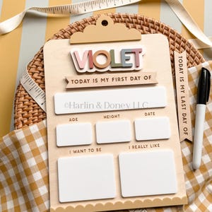 May include: A light-colored wooden clipboard sign with the name VIOLET in raised letters. It features sections for Today is my first day of, Age, Height, Date, I want to be, and I really like. A black marker and a measuring tape are included.