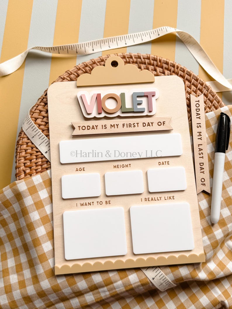 May include: A light-colored wooden clipboard sign with the name VIOLET in raised letters. It features sections for Today is my first day of, Age, Height, Date, I want to be, and I really like. A black marker and a measuring tape are included.