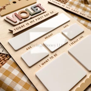 May include: A wooden milestone board featuring the name VIOLET in raised, multi-colored letters. The board includes text sections for Today Is My First Day Of, Today Is My Last Day Of, Date, Height, Age, I Really Like, and I Want To Be.