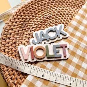 May include: Two layered name signs featuring the names JACK and VIOLET in raised block lettering. The JACK sign has grey letters on a white base, while the VIOLET sign has multi-colored letters in pink, tan, green, and grey on a white base.
