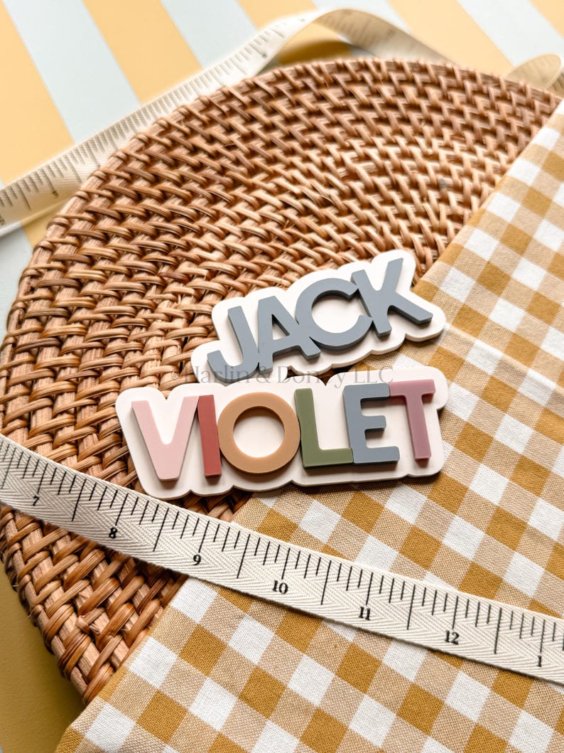 May include: Two layered name signs featuring the names JACK and VIOLET in raised block lettering. The JACK sign has grey letters on a white base, while the VIOLET sign has multi-colored letters in pink, tan, green, and grey on a white base.
