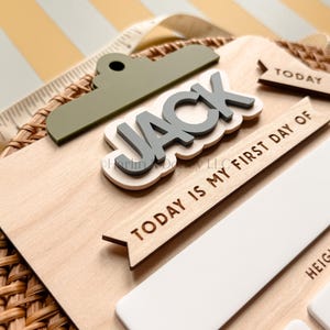 May include: A wooden milestone board featuring a sage green clipboard-style top, the name JACK in raised grey letters, and the text TODAY IS MY FIRST DAY OF. The board includes white rectangular sections for details like AGE and HEIGHT.