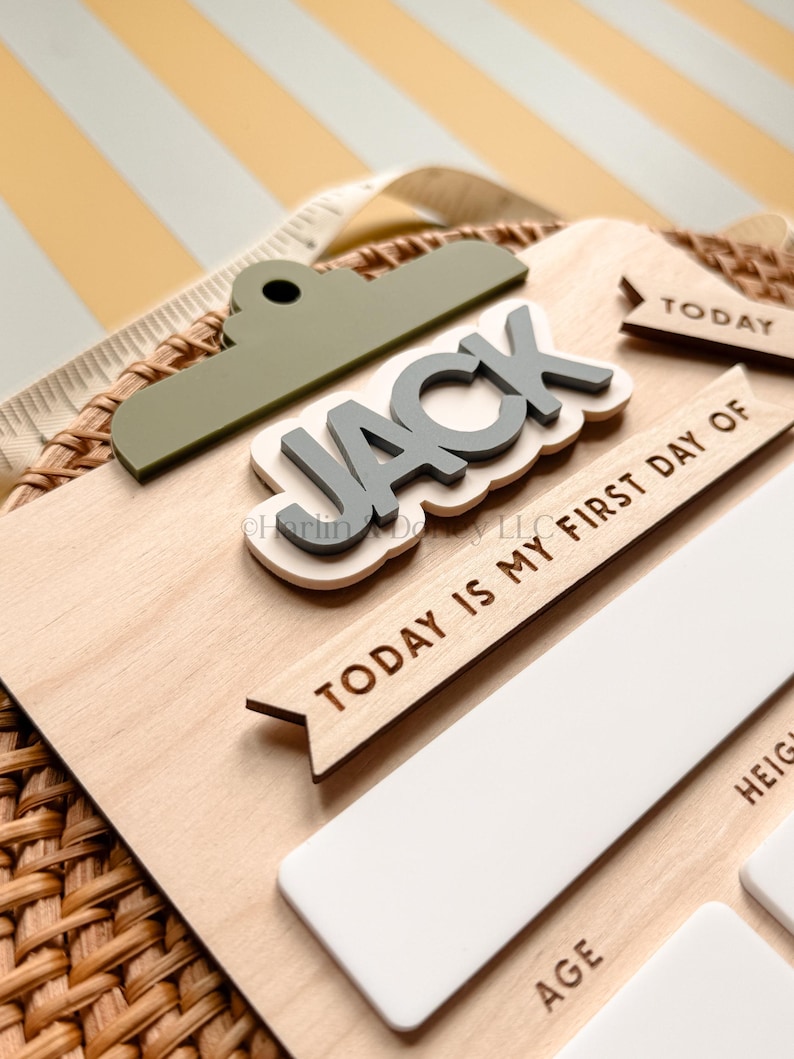 May include: A wooden milestone board featuring a sage green clipboard-style top, the name JACK in raised grey letters, and the text TODAY IS MY FIRST DAY OF. The board includes white rectangular sections for details like AGE and HEIGHT.