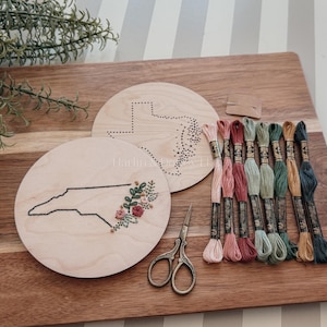 May include: A wooden embroidery kit featuring a North Carolina state outline with floral accents and a Texas outline. Includes embroidery floss in various colors, scissors, and a needle. The kit is displayed on a wooden cutting board.