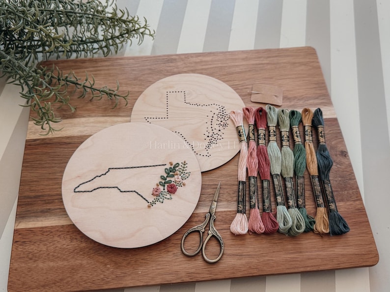 May include: A wooden embroidery kit featuring a North Carolina state outline with floral accents and a Texas outline. Includes embroidery floss in various colors, scissors, and a needle. The kit is displayed on a wooden cutting board.