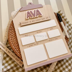 May include: A light wood clipboard sign with a mauve top clip and the name AVA in raised letters. It features the text TODAY IS MY FIRST DAY OF and blank white rectangular spaces for age, height, date, likes, and goals, with a black marker.