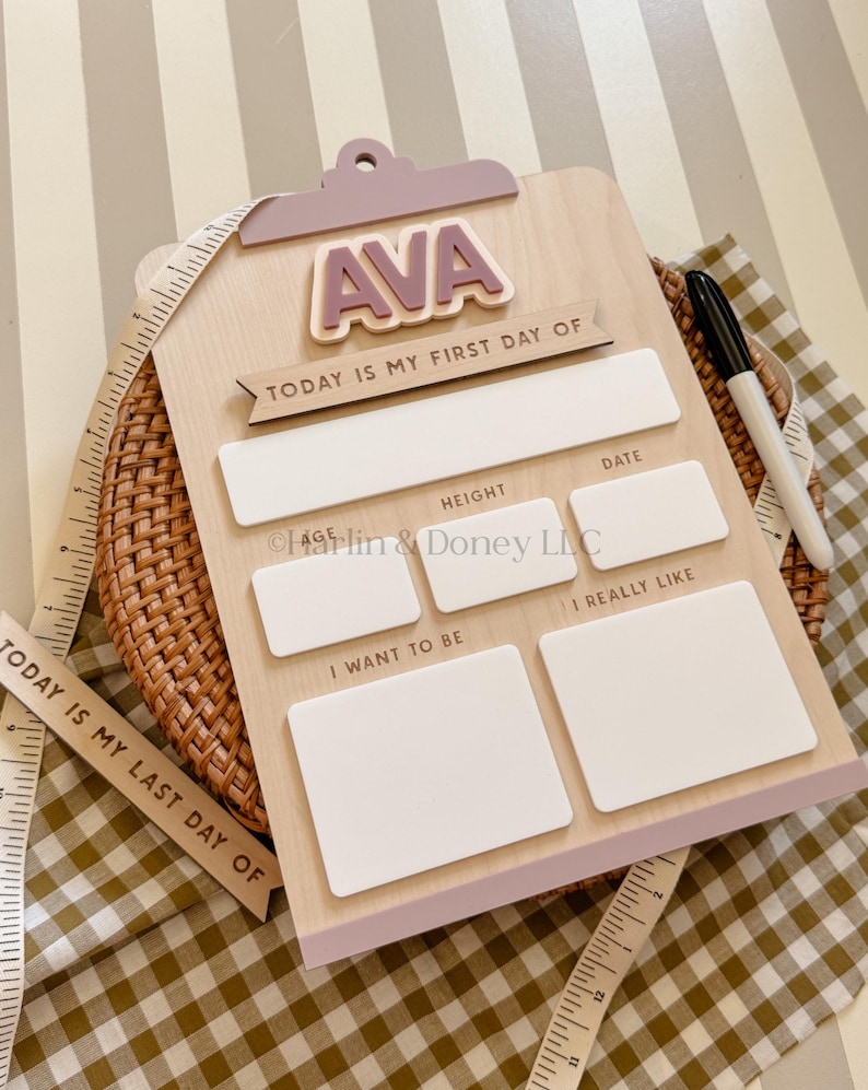 May include: A light wood clipboard sign with a mauve top clip and the name AVA in raised letters. It features the text TODAY IS MY FIRST DAY OF and blank white rectangular spaces for age, height, date, likes, and goals, with a black marker.