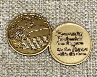 May include: Two gold-toned metal tokens. One side features a scenic image of a lake with trees and a cross. The other side has the text "Serenity isn't freedom from the storm it's the Peace within the storm".