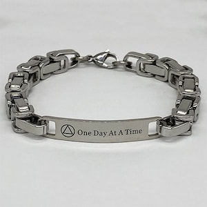 Può includere: Un braccialetto d'argento con un design a maglie di catena e un'etichetta rettangolare che recita "One Day At A Time". L'etichetta presenta anche un simbolo triangolare.