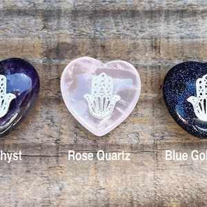 May include: Three heart-shaped crystal stones with a white Hamsa hand design. The stones are amethyst, rose quartz, and blue goldstone.