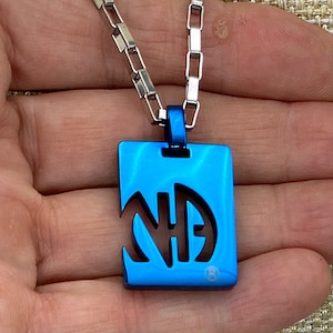 Puede incluir: Un collar con un colgante de metal azul y una cadena plateada. El colgante es rectangular y tiene las letras "NA" recortadas.