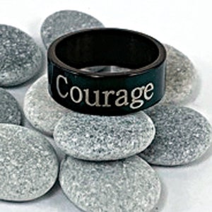 Könnte beinhalten: Ein schwarzer Ring mit der Gravur "Courage". Der Ring liegt auf einem Stapel glatter, grauer Steine.