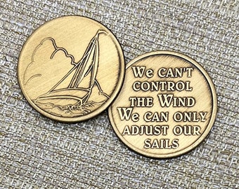 May include: Two gold-toned metal tokens. One token features a sailboat on the water with a cloudy sky. The other token has the text "We can't control the wind. We can only adjust our sails."