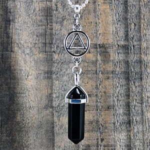 Puede incluir: Collar plateado con un colgante de cristal negro hexagonal. El colgante está rematado en plata y suspendido de una cadena. Incluye un símbolo triangular en un círculo. Fondo de madera envejecida.