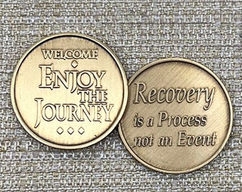 May include: Two bronze-colored challenge coins. One coin reads "WELCOME ENJOY THE JOURNEY" and the other reads "Recovery is a Process not an Event". The coins are circular with raised lettering.