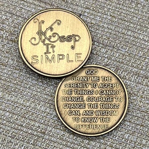 May include: Two gold-colored metal coins. One coin reads "Keep It Simple" in an elegant font. The other coin has an inscription that reads "God grant me the serenity to accept the things I cannot change..." The coins are on a textured surface.