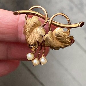 May include: A gold-toned brooch featuring a leaf design with three small, dangling pearls. The brooch has a curved, flowing shape with detailed leaf textures and a polished finish, suitable for adding a touch of elegance.