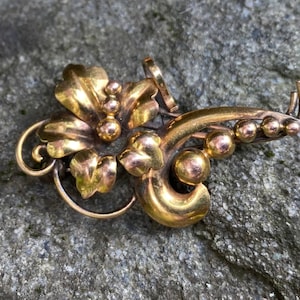 May include: A gold-toned brooch with a floral design. The brooch features a central flower with leaves and a series of small, round beads.