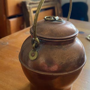 May include: A small, antique copper kettle with a brass handle and lid. The kettle has a rounded body and a small, circular lid. The handle arches over the top, and the kettle sits on a wooden surface. The copper has a warm, reddish-brown hue.