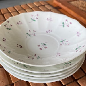 May include: A set of four white ceramic saucers with a delicate floral pattern in shades of purple and green.