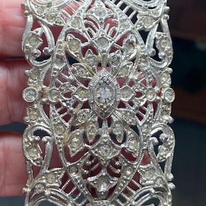 May include: Silver-toned rectangular brooch with an ornate filigree design. The brooch is encrusted with numerous small, clear rhinestones, creating a sparkling effect. The intricate metalwork features floral and geometric patterns.