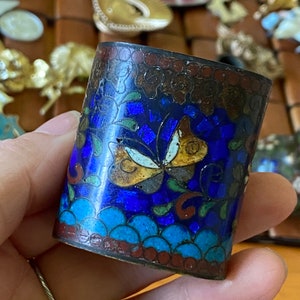 May include: A small, round, blue and gold enamel box with a butterfly design. The box has a red and brown border.